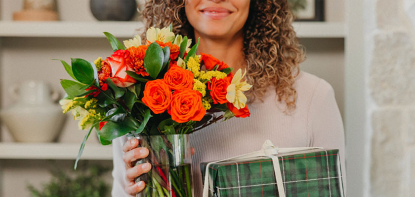 A cheerful woman holding a vibrant bouquet of orange roses and a green gift box.