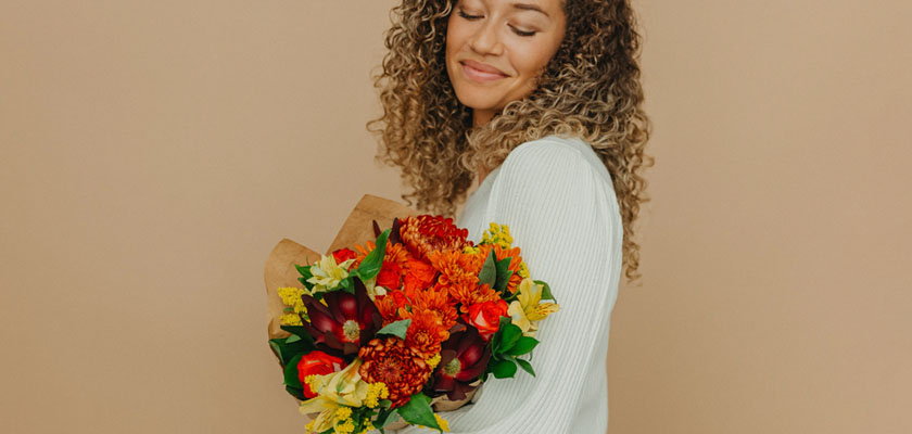A cheerful woman holds a vibrant floral bouquet featuring warm autumn hues and rich textures.