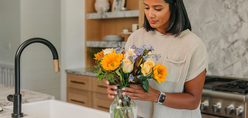 A woman carefully arranges vibrant sunflowers and roses in a glass vase on a kitchen countertop.