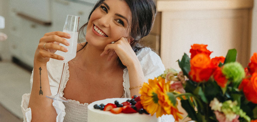 A smiling woman holds a drink near a colorful floral arrangement and a delicious cake.