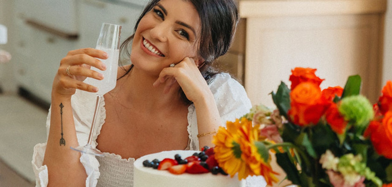 A smiling woman holds a drink near a colorful floral arrangement and a delicious cake.