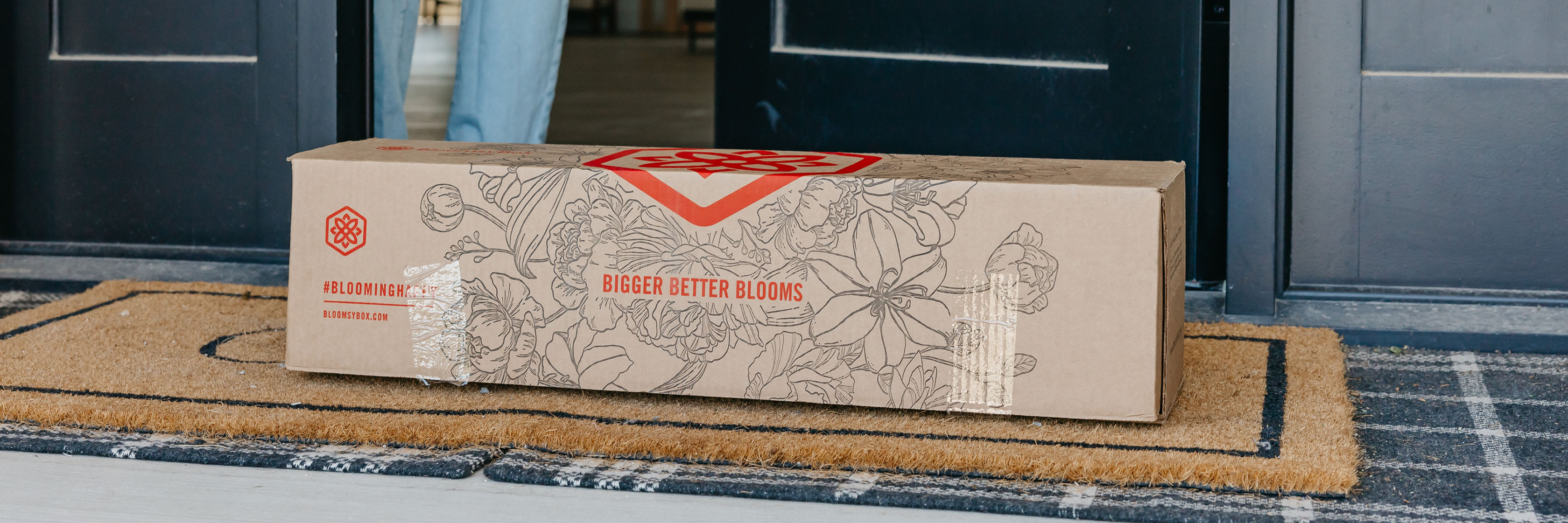 A beautifully packaged floral delivery sitting on a welcome mat at a front door.
