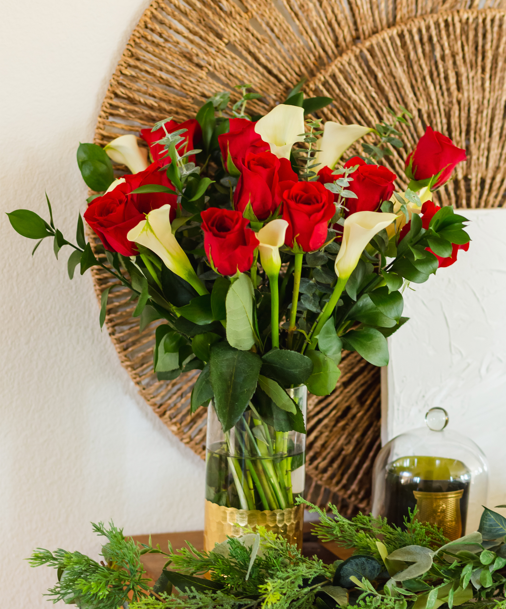 Vibrant red roses and white calla lilies arranged in a chic glass vase, surrounded by lush greenery.