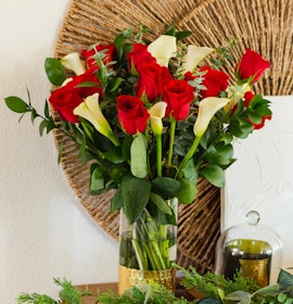 Vibrant red roses and white calla lilies arranged in a chic glass vase, surrounded by lush greenery.