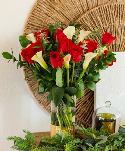 Vibrant red roses and white calla lilies arranged in a chic glass vase, surrounded by lush greenery.