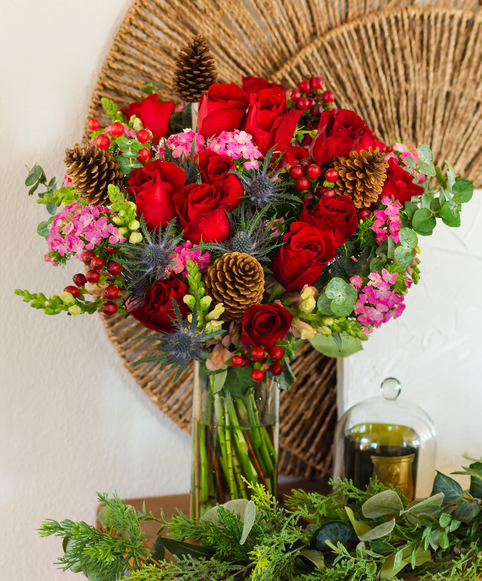 A vibrant bouquet featuring red roses, pink flowers, pinecones, and lush greenery in a glass vase.