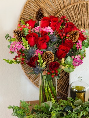 Vibrant bouquet featuring red roses, pink flowers, and pinecones artfully arranged in a glass vase.