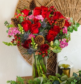 Vibrant bouquet featuring red roses, pink flowers, and pinecones artfully arranged in a glass vase.