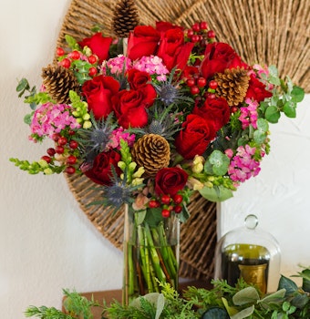 Vibrant bouquet featuring red roses, pink flowers, and pinecones artfully arranged in a glass vase.