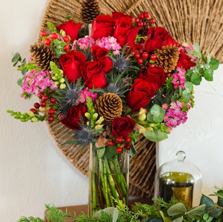 Vibrant bouquet featuring red roses, pink flowers, and pinecones artfully arranged in a glass vase.