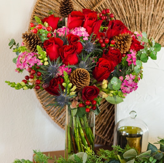 Vibrant bouquet featuring red roses, pink flowers, and pinecones artfully arranged in a glass vase.