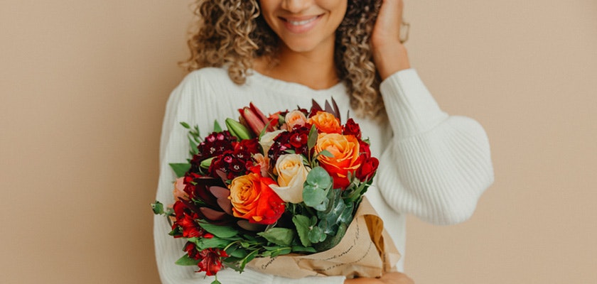 A cheerful woman holding a vibrant bouquet of orange and red flowers, showcasing seasonal blooms.