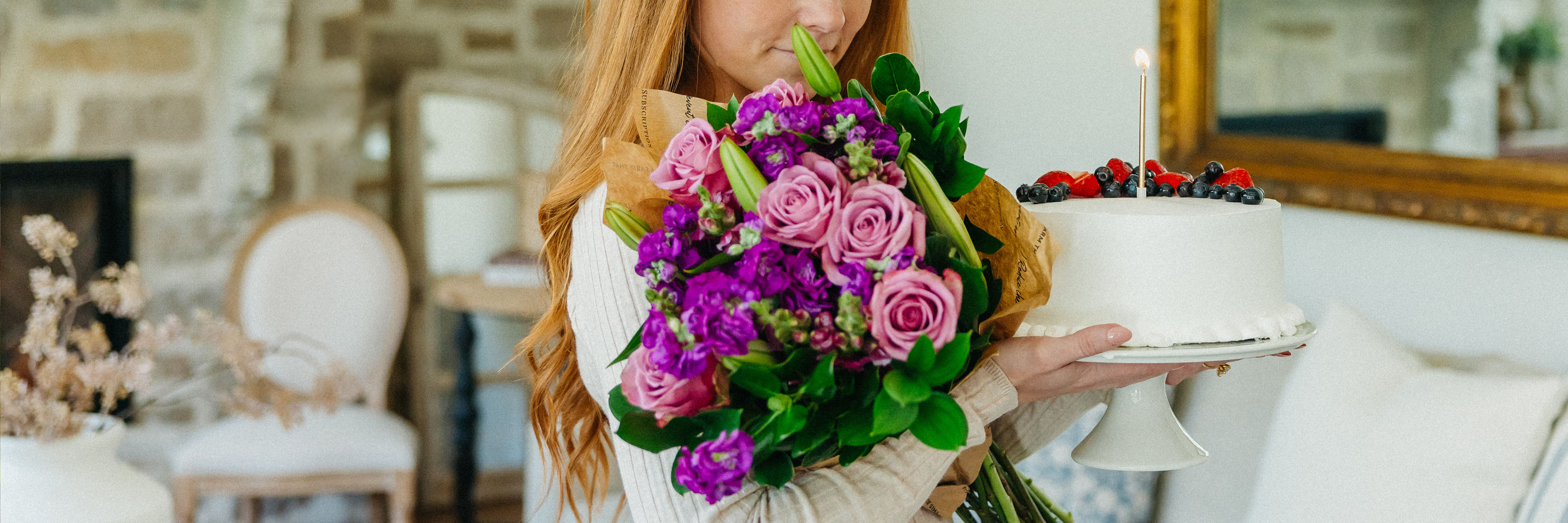 Woman holding a vibrant bouquet of purple roses and a cake topped with berries in a cozy setting.