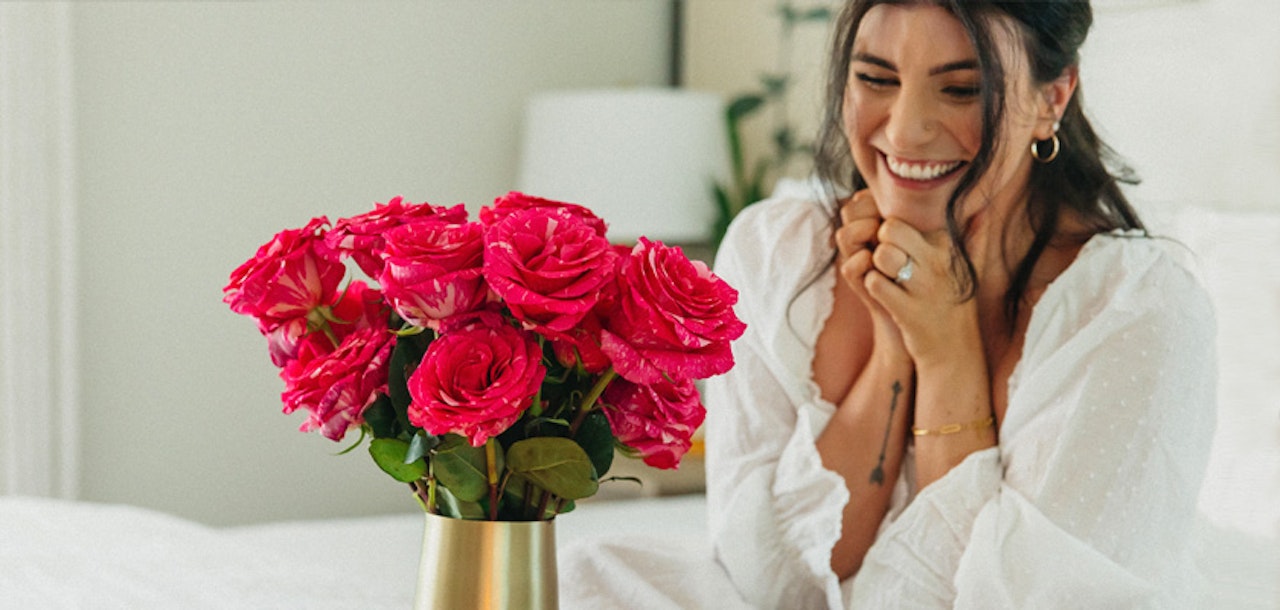 A cheerful woman smiles joyfully beside a vibrant pink rose bouquet in a golden vase.
