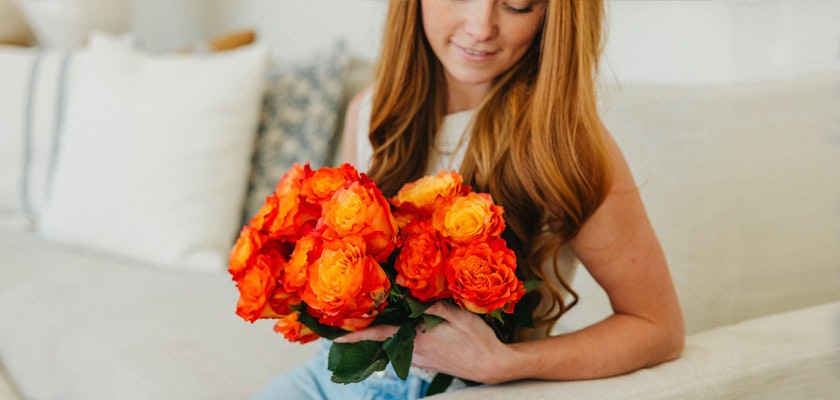 A lovely woman holding a vibrant bouquet of orange and yellow roses, sitting on a cozy couch.