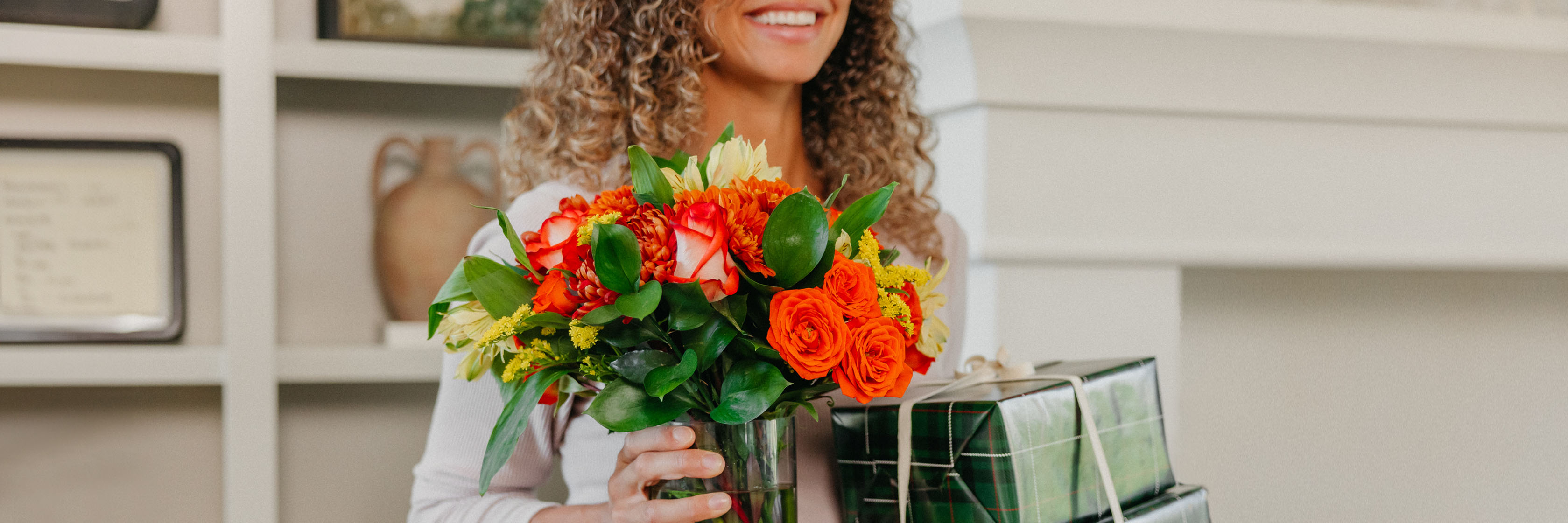 A cheerful woman holding a vibrant bouquet of red and orange flowers alongside wrapped gifts.