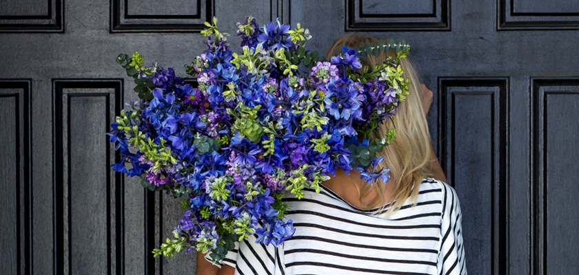 A woman holds a vibrant bouquet of blue and purple flowers against a stylish wooden backdrop.