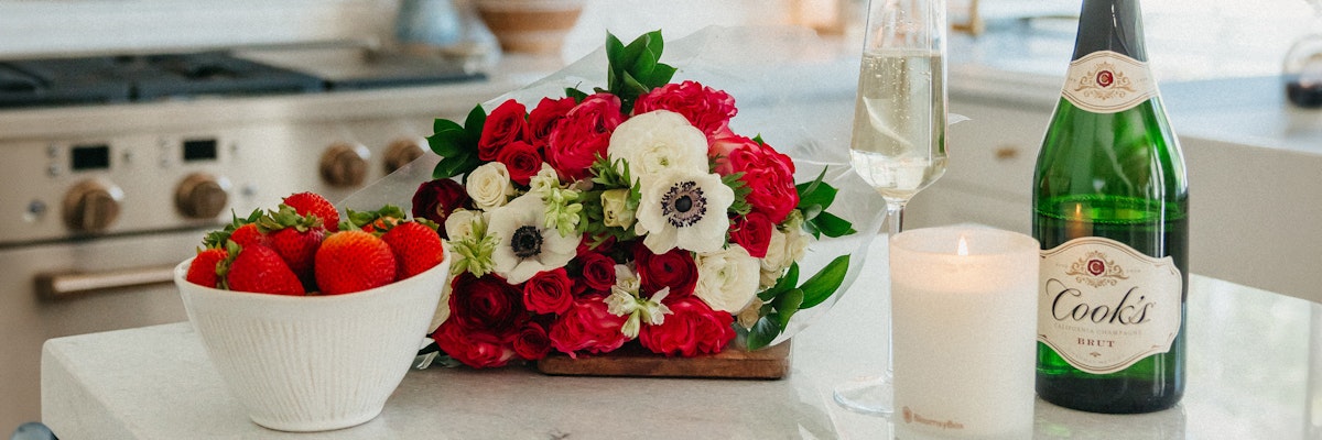 Fresh red and white floral arrangement beside strawberries and sparkling wine on a kitchen counter.