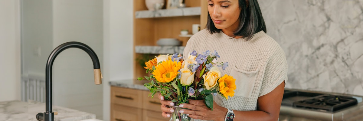 A woman holding a vibrant bouquet of sunflowers and pastel flowers in a modern kitchen setting.