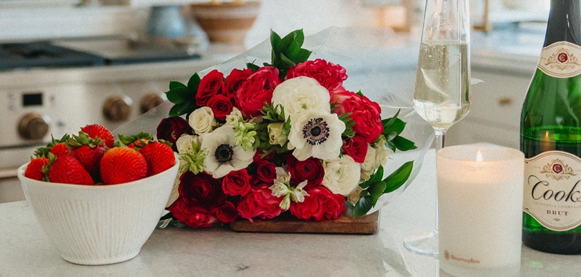 A vibrant bouquet of red roses and white anemones beside strawberries and champagne.