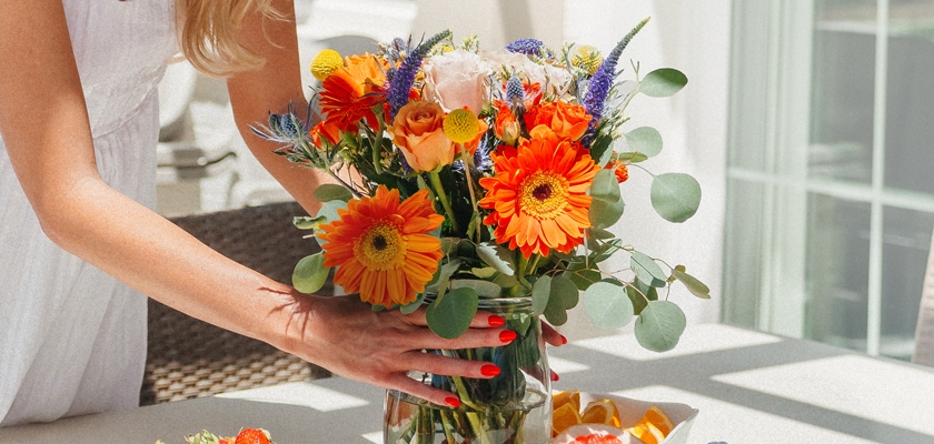 A vibrant floral arrangement featuring orange gerbera daisies, roses, and greenery on a sunny table.