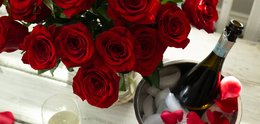 Lush red roses in a vase beside a champagne bottle and petals, ideal for romantic celebrations.