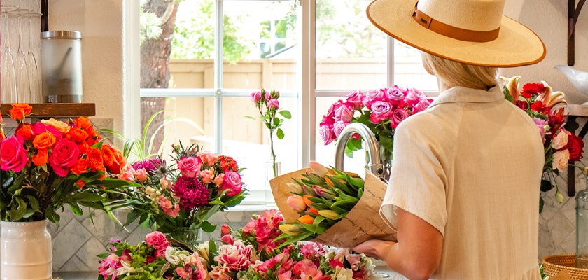 A woman in a large hat arranging vibrant flowers in a bright kitchen filled with colorful blooms.