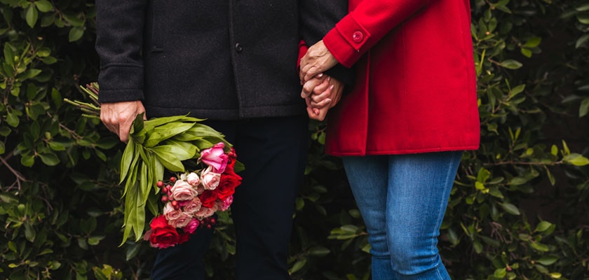 Couple holding hands while one person carries a vibrant bouquet of roses amidst lush greenery.