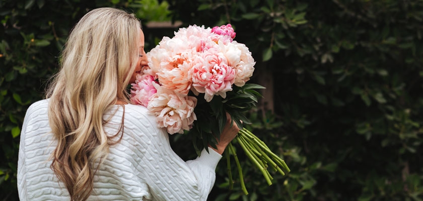 A woman holding a lush bouquet of pink peonies, surrounded by greenery, in a serene outdoor setting.