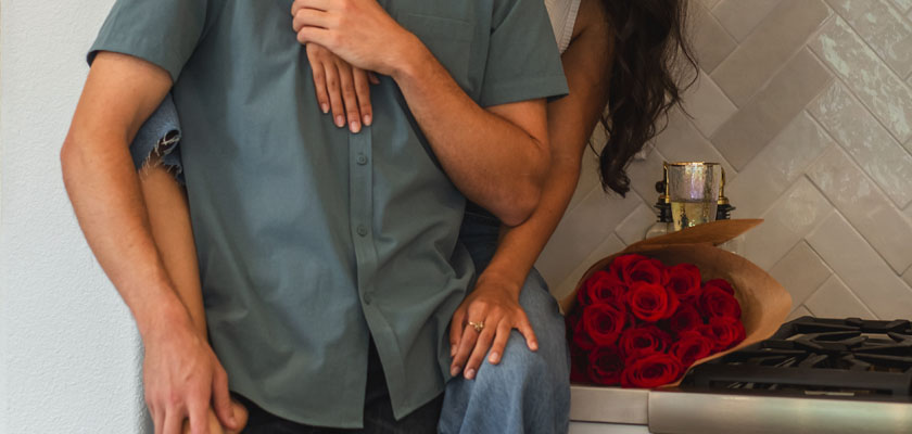 A couple sharing a cozy moment in a kitchen with a vibrant bouquet of red roses nearby.