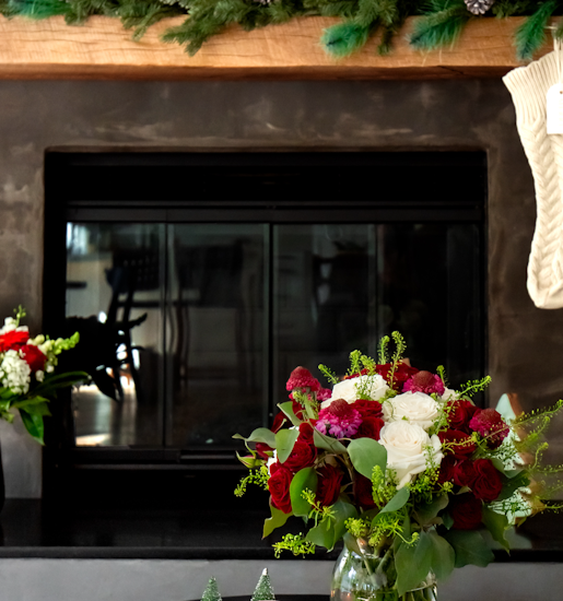 A festive winter bouquet featuring red roses and seasonal greens by a cozy fireplace.