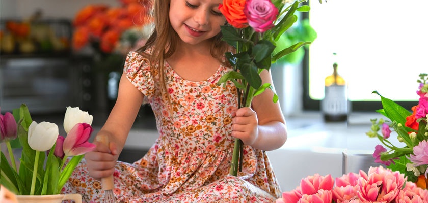 A cheerful girl in a floral dress arranging vibrant flowers in a bright kitchen.