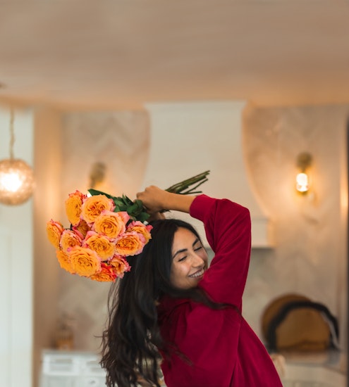 A joyful woman in a red top holding a vibrant bouquet of orange roses, celebrating in a bright room.