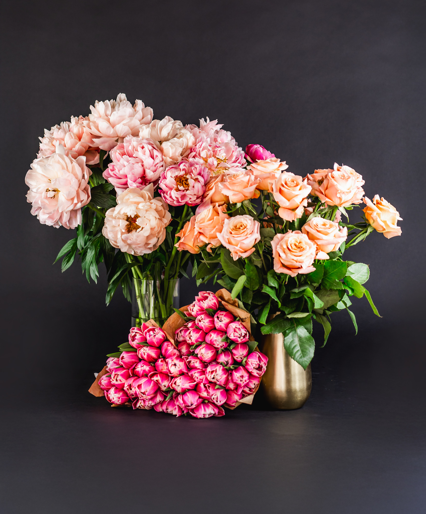 A vibrant arrangement of peonies and peach roses in elegant vases against a dark background.
