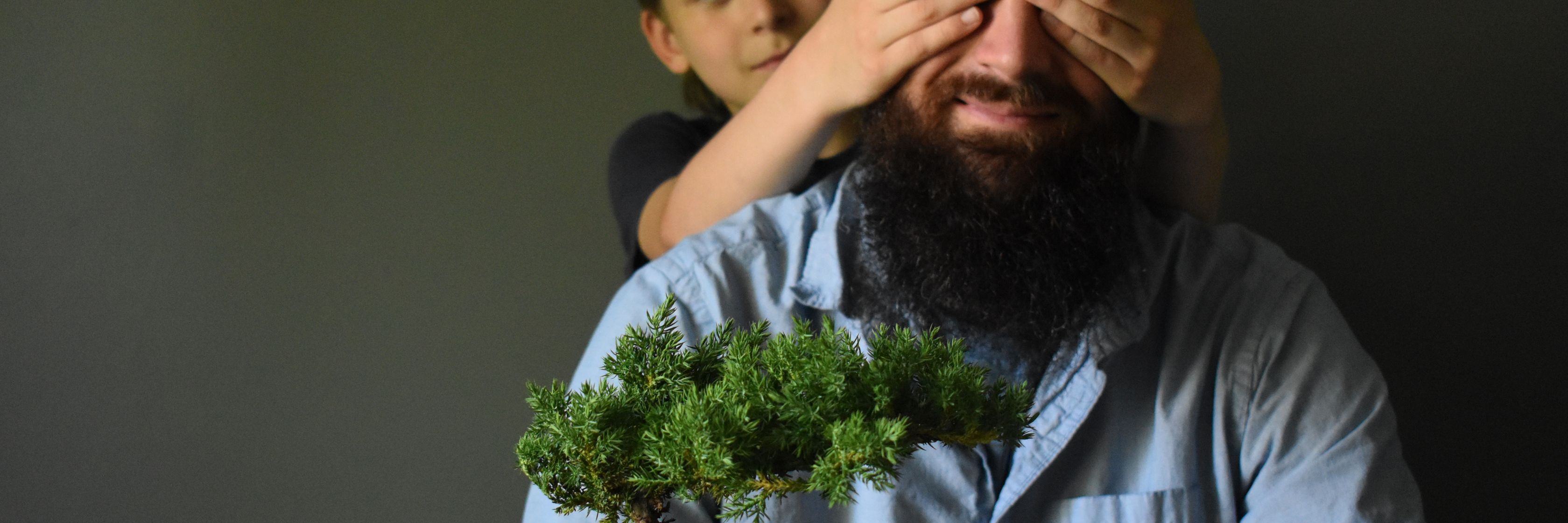 A joyful moment capturing a child playfully covering a man's eyes while he holds a bonsai tree.
