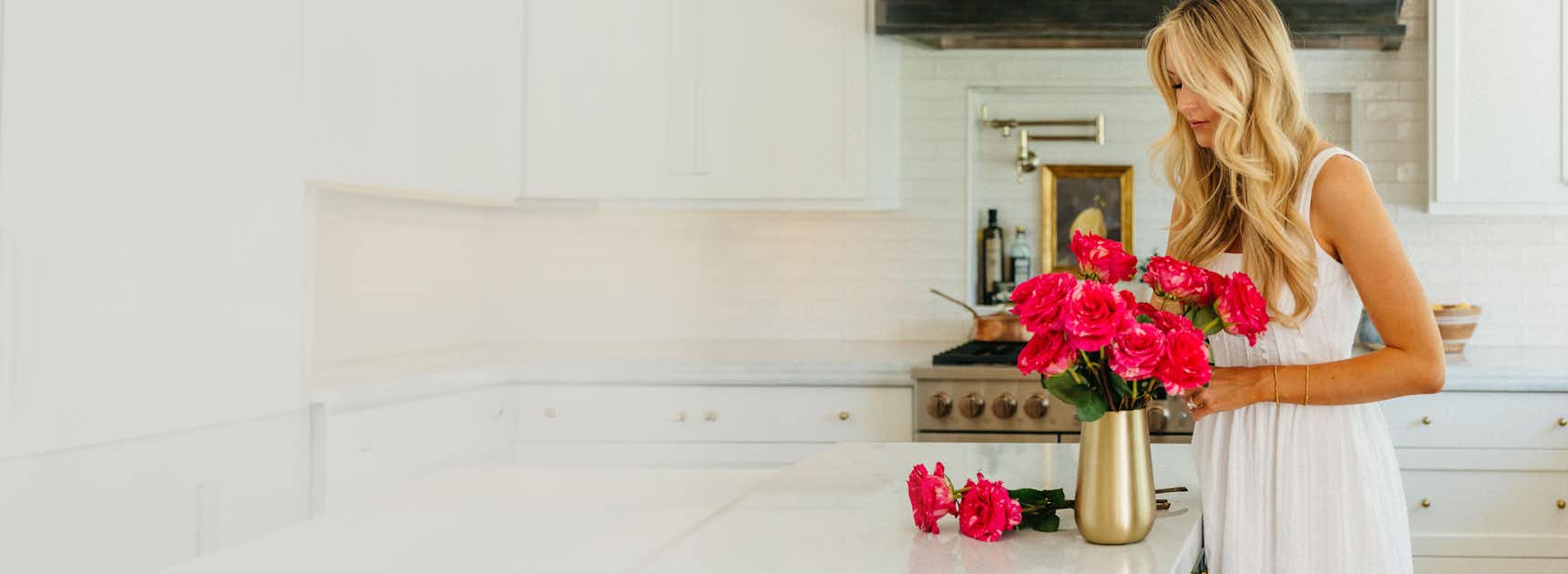 A woman in a white dress arranges vibrant pink roses in a gold vase on a kitchen counter.