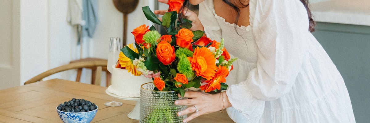 A woman in a white dress joyfully arranges a vibrant floral bouquet with orange and yellow blooms.