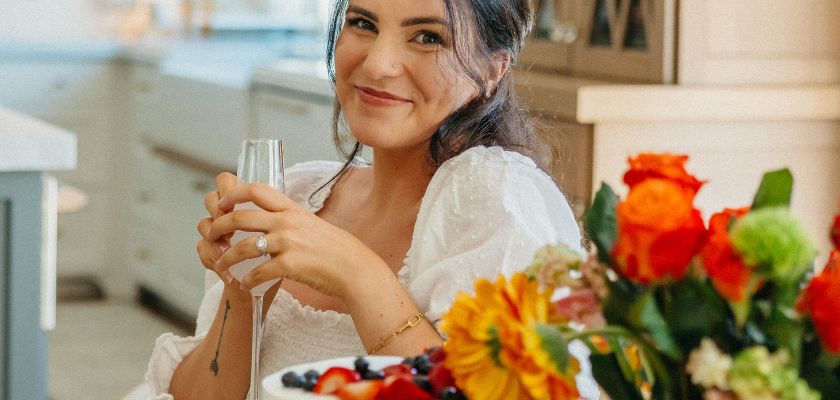 A joyful woman enjoying a drink, surrounded by a vibrant fruit platter and colorful flowers.