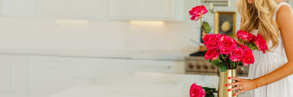 A woman in a white dress arranging vibrant pink roses in a chic gold vase.