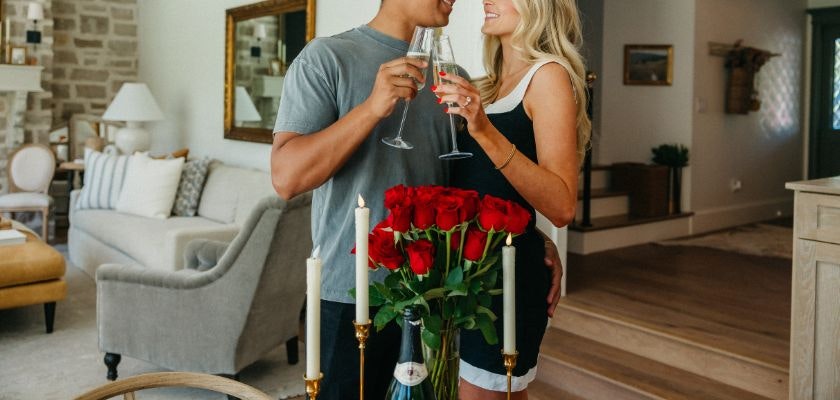 Couple celebrating with champagne in a cozy home setting, surrounded by red roses.