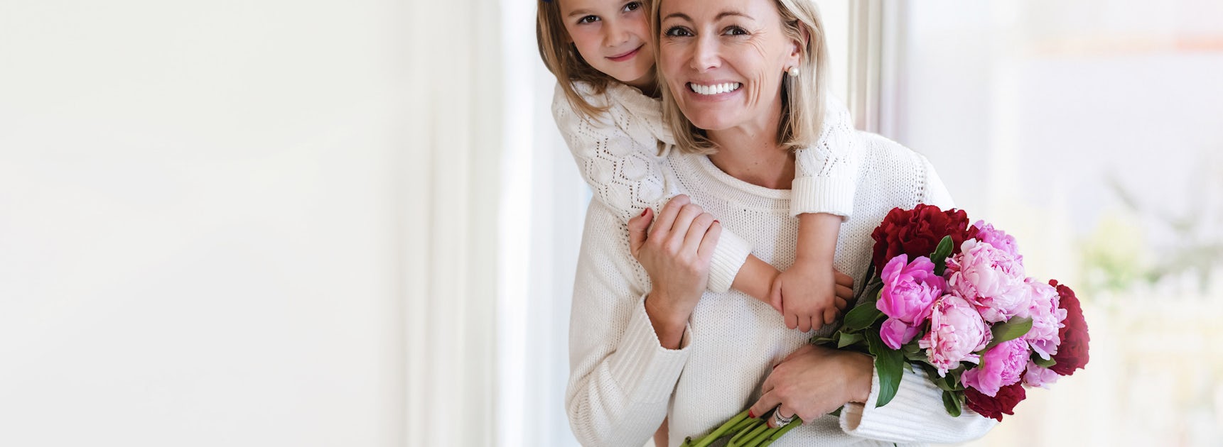 A joyful woman holding a colorful bouquet, smiling with her daughter embracing her from behind.