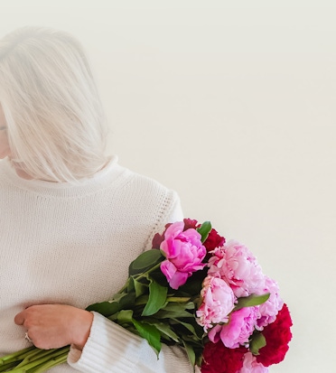 A woman in a cozy sweater holding a vibrant bouquet of pink and red peonies.