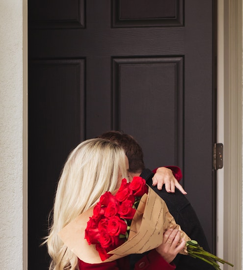A couple shares a romantic kiss at the door, with a bouquet of vibrant red roses.