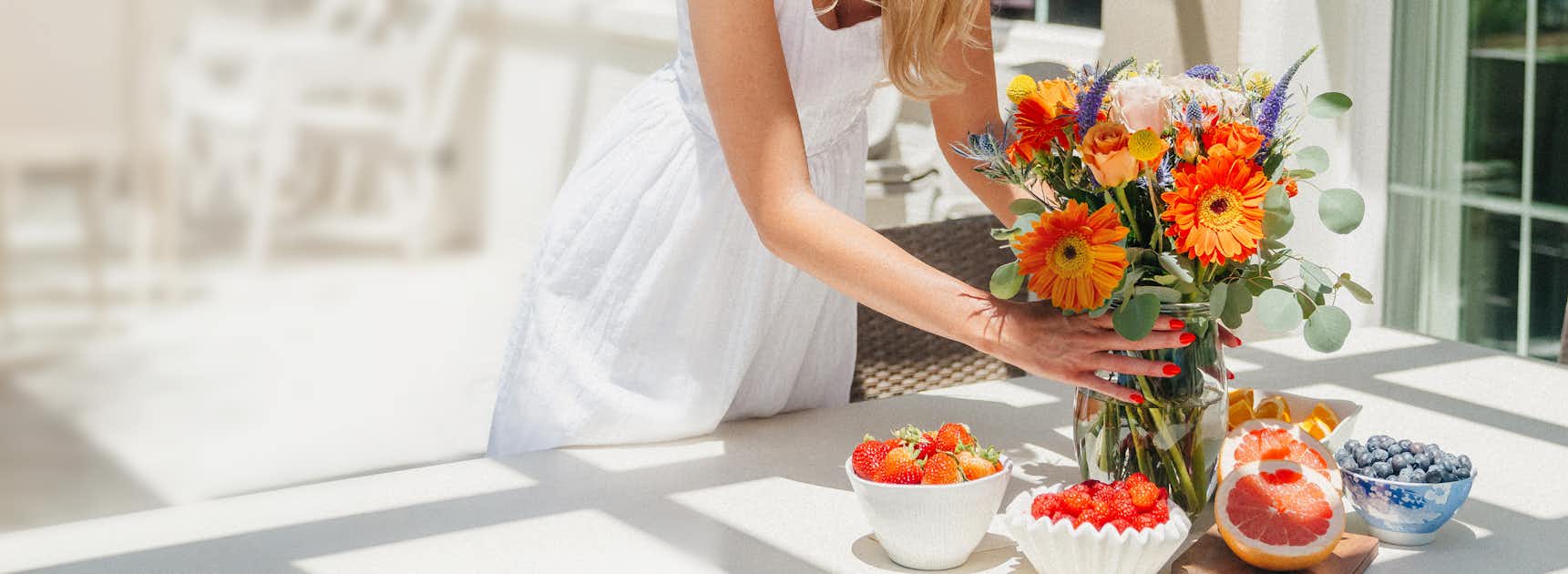 A woman arranging a vibrant bouquet of daisies and gerberas on a sunlit table with fresh fruits.