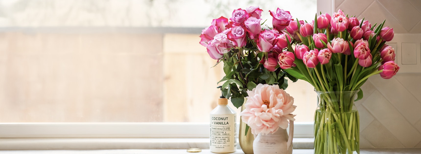 Lush pink tulips and roses arranged in elegant vases on a sunlit windowsill.