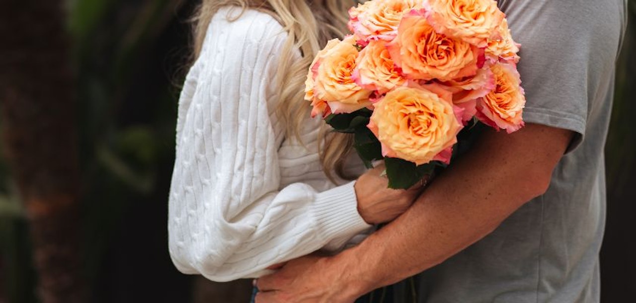 A couple embracing, with a stunning bouquet of peach and pink roses in hand, showcasing love and joy.
