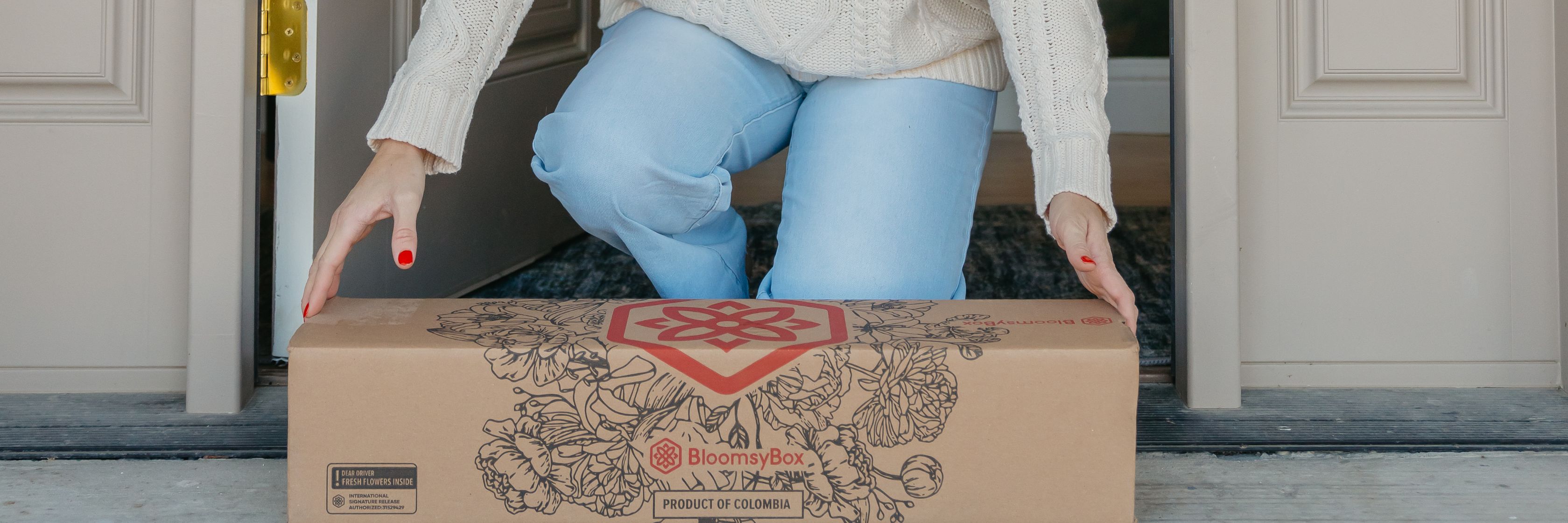 A person in a cozy sweater receiving a BloomsyBox floral delivery at home.