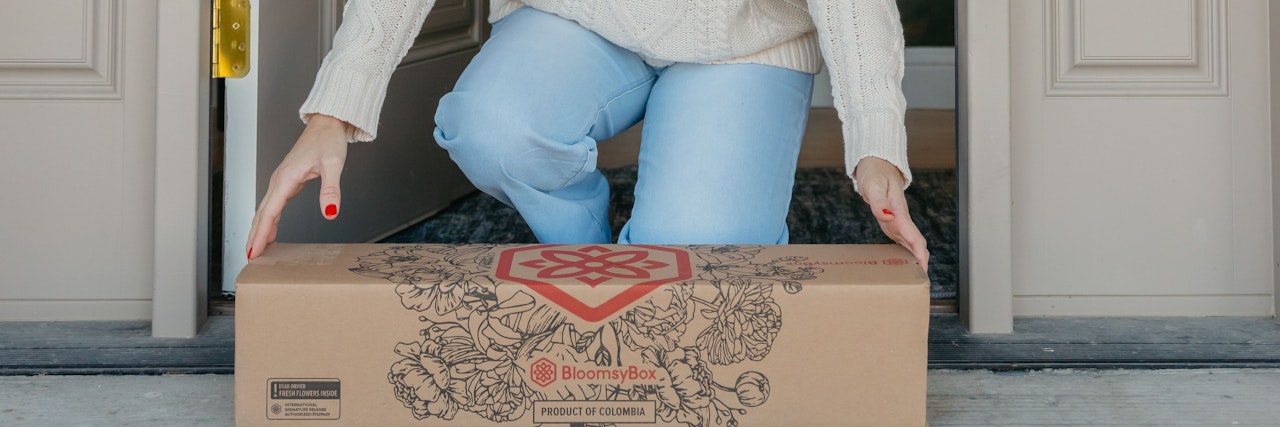 A person in a cozy sweater receiving a BloomsyBox floral delivery at home.