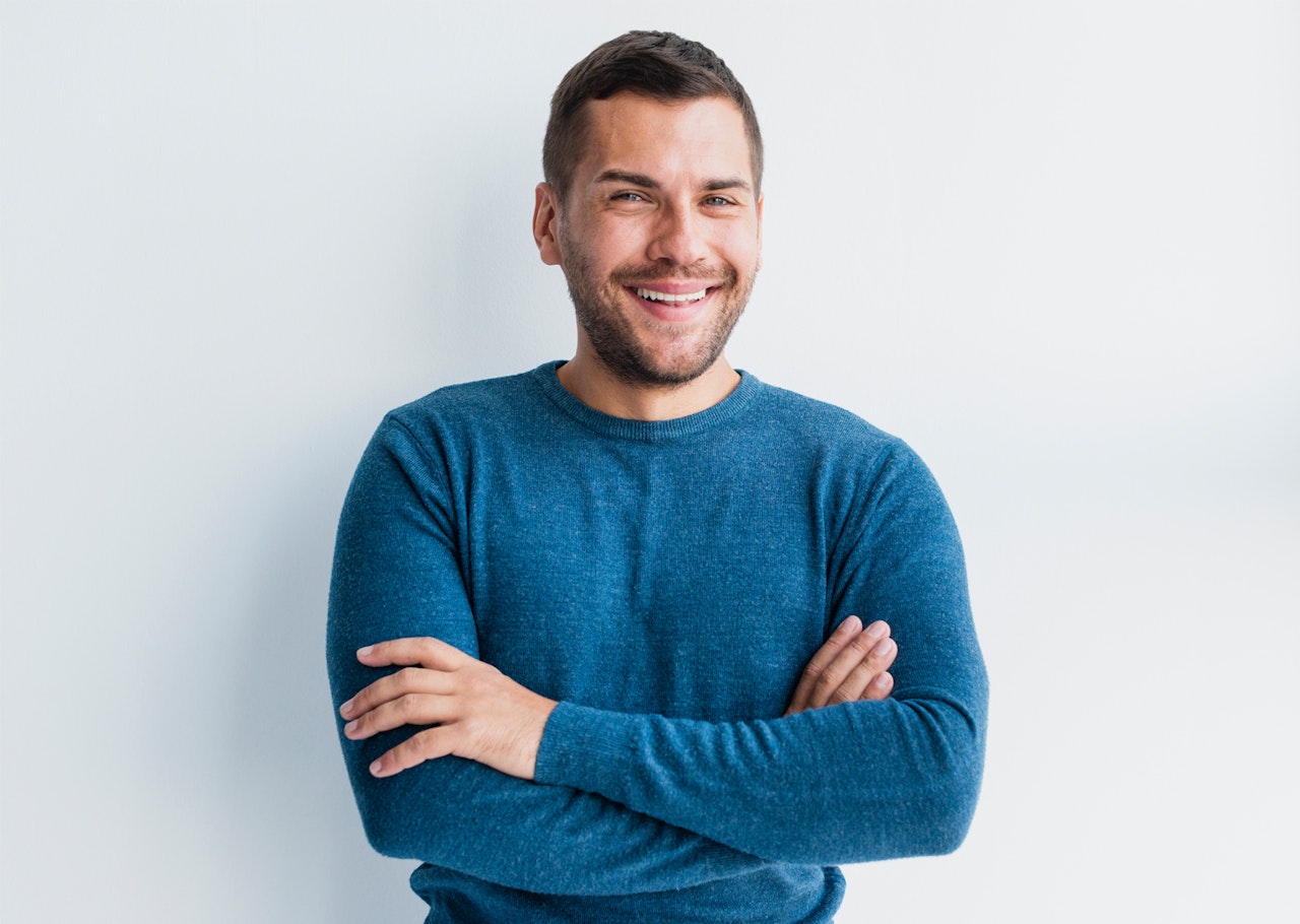 A cheerful young man in a blue sweater smiles confidently with crossed arms against a light background.