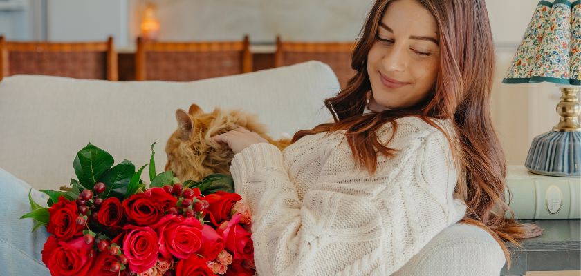 A woman in a cozy sweater enjoys a vibrant bouquet of red roses while sitting with her cat.